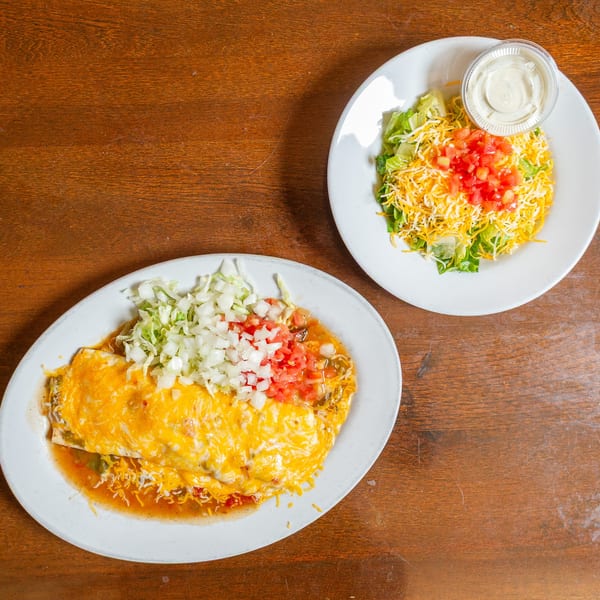 Two white plates of Mexican food on a wooden table, one with a cheesy burrito, the other a salad.
