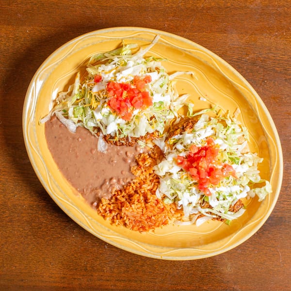 Two tacos with lettuce, cheese, sour cream, and diced tomatoes, served with refried beans and rice.