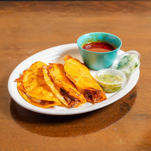 Four crispy birria tacos with a side of consommé, green salsa, and chopped onions on a white plate.
