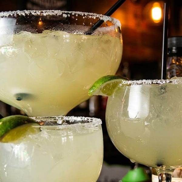 Three large, salt-rimmed margarita glasses filled with icy drinks and lime wedges on a bar.