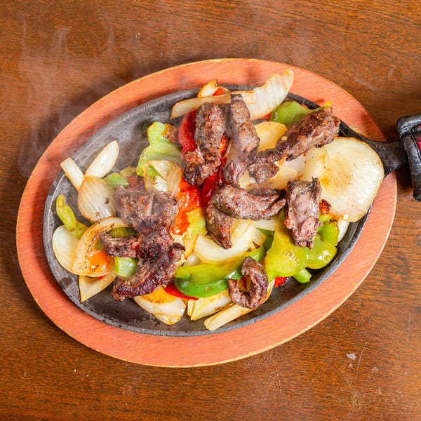 Sizzling steak fajitas with onions and bell peppers on a cast iron skillet with a wooden base.