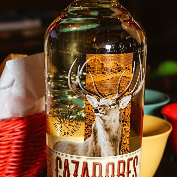 Cazadores tequila bottle with a deer label, next to a basket of tortilla chips and colorful bowls.