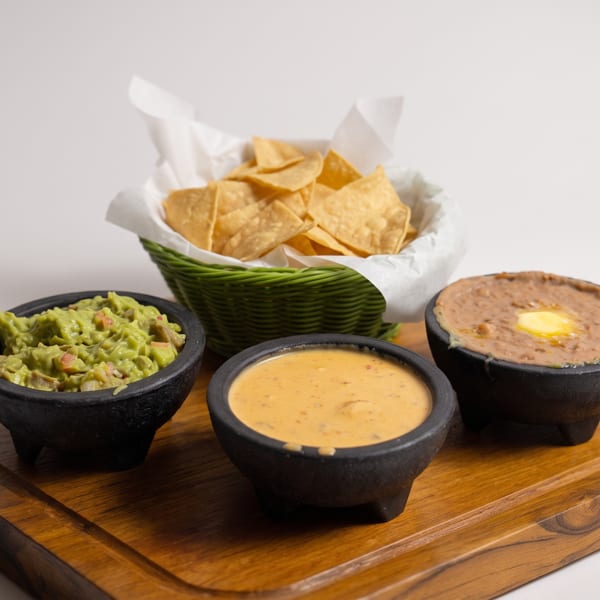 Guacamole, queso, and refried beans in black bowls with a basket of tortilla chips.