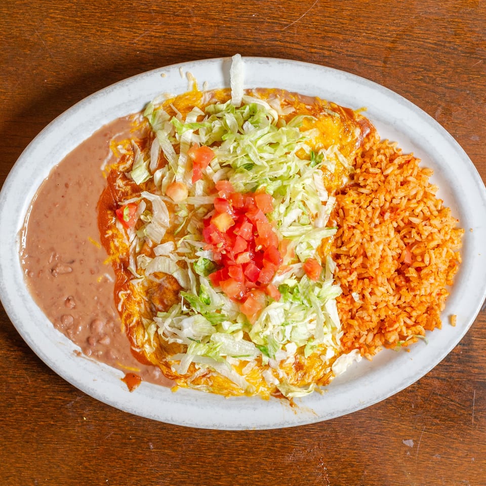 A white oval plate holds a Mexican dish with refried beans, enchiladas topped with lettuce and tomatoes, and orange rice.