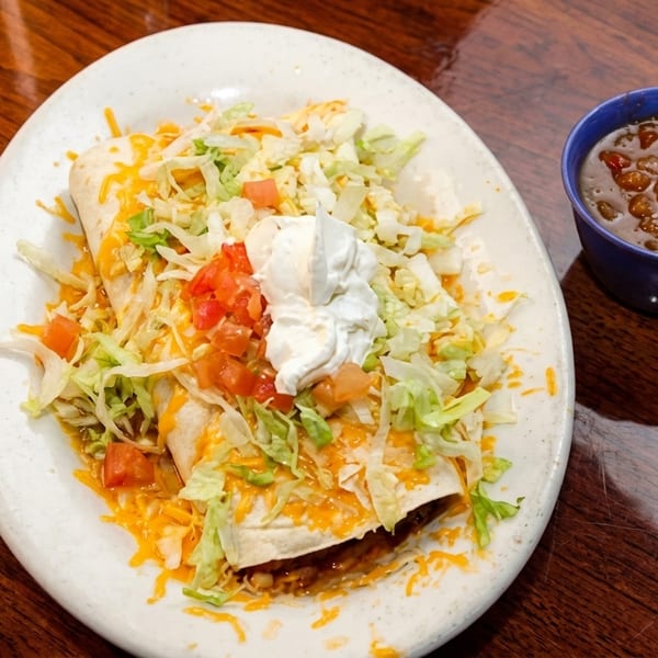 Two burritos topped with shredded lettuce, cheese, diced tomatoes, and sour cream, with a side of salsa.