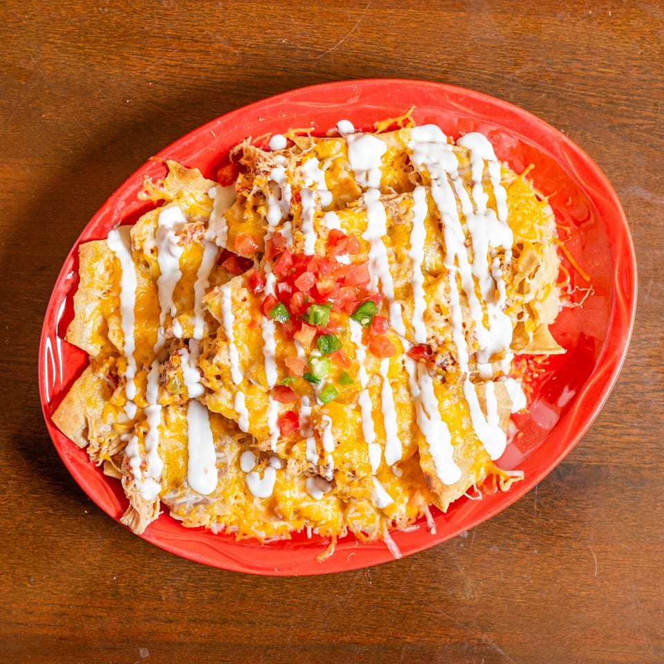A red oval plate filled with cheesy nachos, topped with sour cream, diced tomatoes, and green peppers.