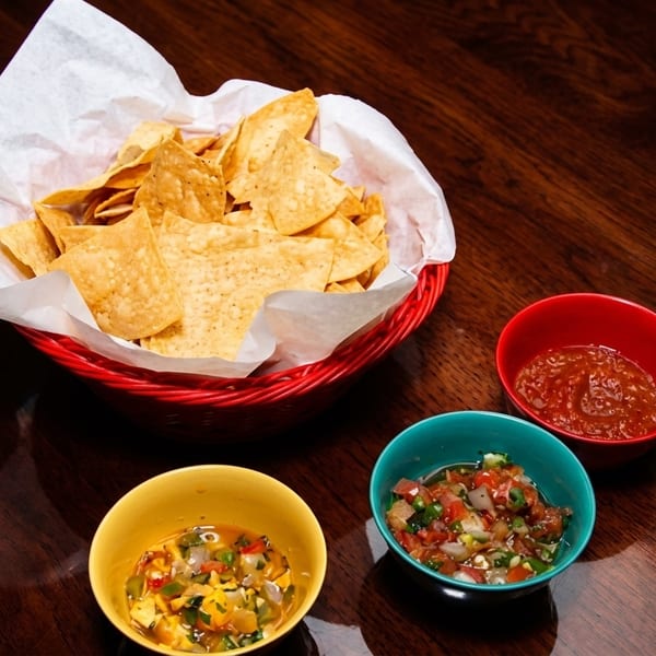 A red basket of tortilla chips with three colorful bowls of salsa and queso on a wooden table.
