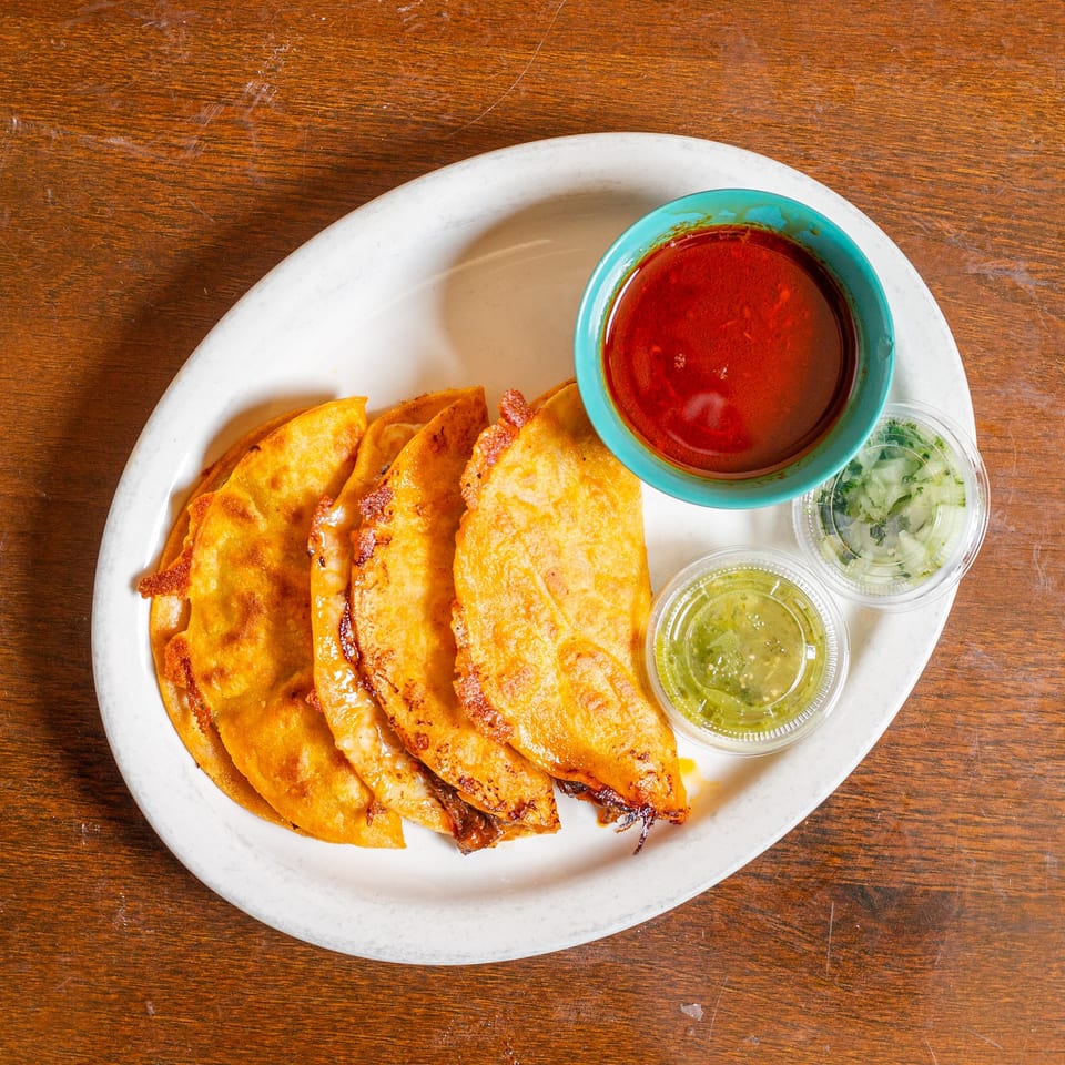Four crispy birria tacos with a side of consommé, green salsa, and diced onions on a white plate.